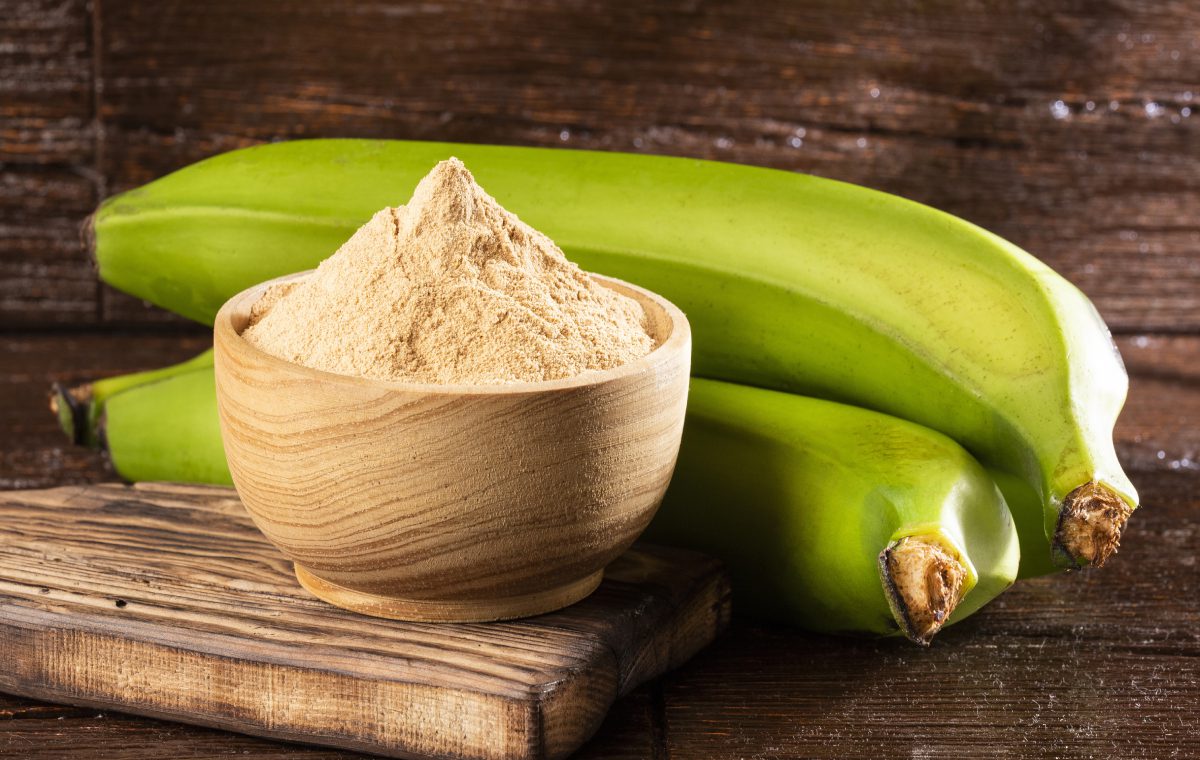 A wooden bowl of powdered milk next to two green bananas.